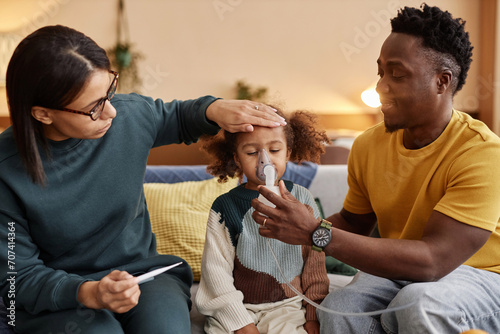 Medium long shot of family at home, mom measuring African American daughters temperature while father holding nebulizer mask at childs face photo