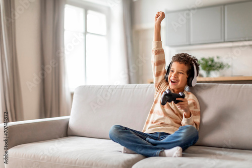 Little black boy in headphones holding joystick and raising hand in victory photo