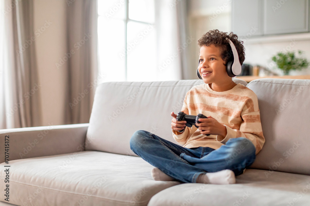 Obraz premium Engrossed black little boy in headphones using game controller at home