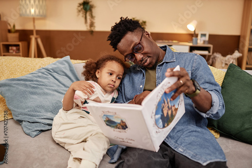 Medium long shot of African American dad wearing glasses reading fairy tales to sleepy little daughter while sitting on couch photo