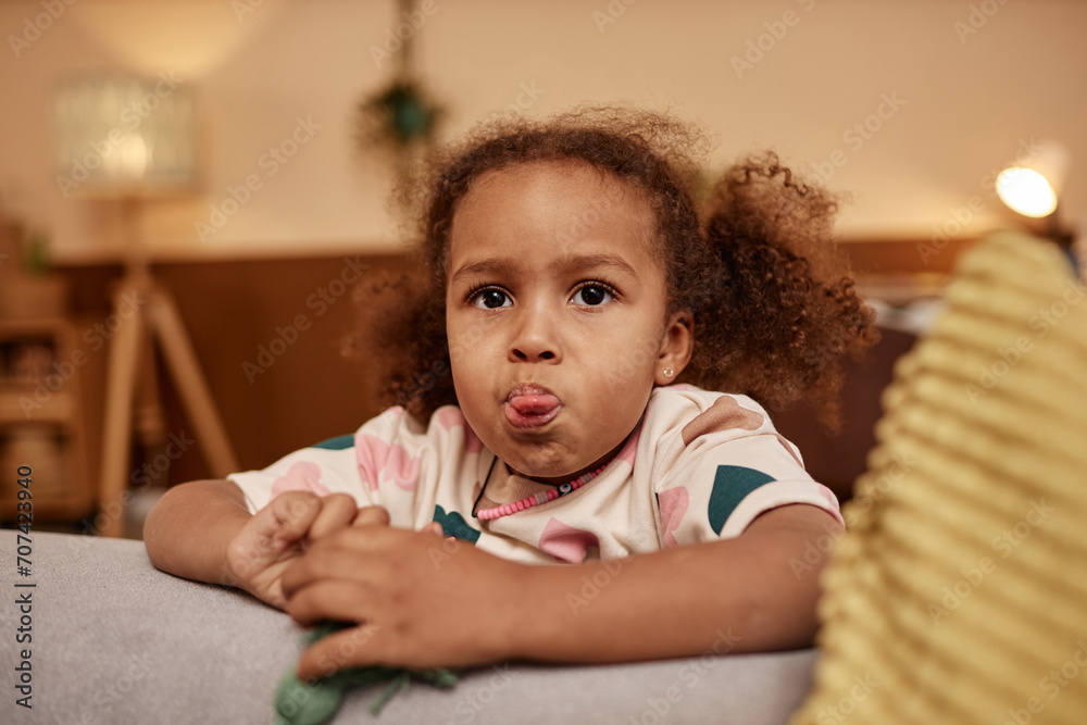 Naklejka premium Medium close up shot of African American preschool girl with curly ponytails sticking out her tongue while leaning on couch