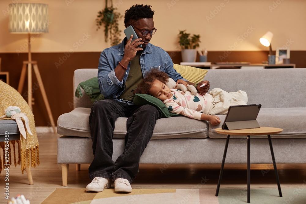 Full shot of Black man with thermometer in hand calling ambulance using smartphone for his preschool daughter lying on sofa in living room