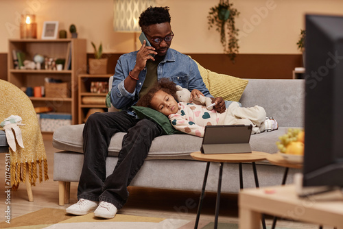 Wide shot of sick African American girl lying on couch and hugging toy looking at tablet while her concerned father talking on phone with pediatrician photo