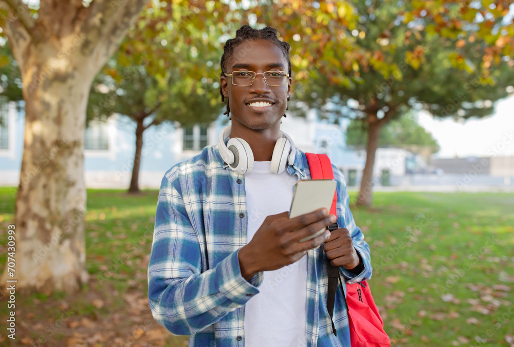 Fototapeta premium African American guy standing with phone, headphones and backpack outdoors