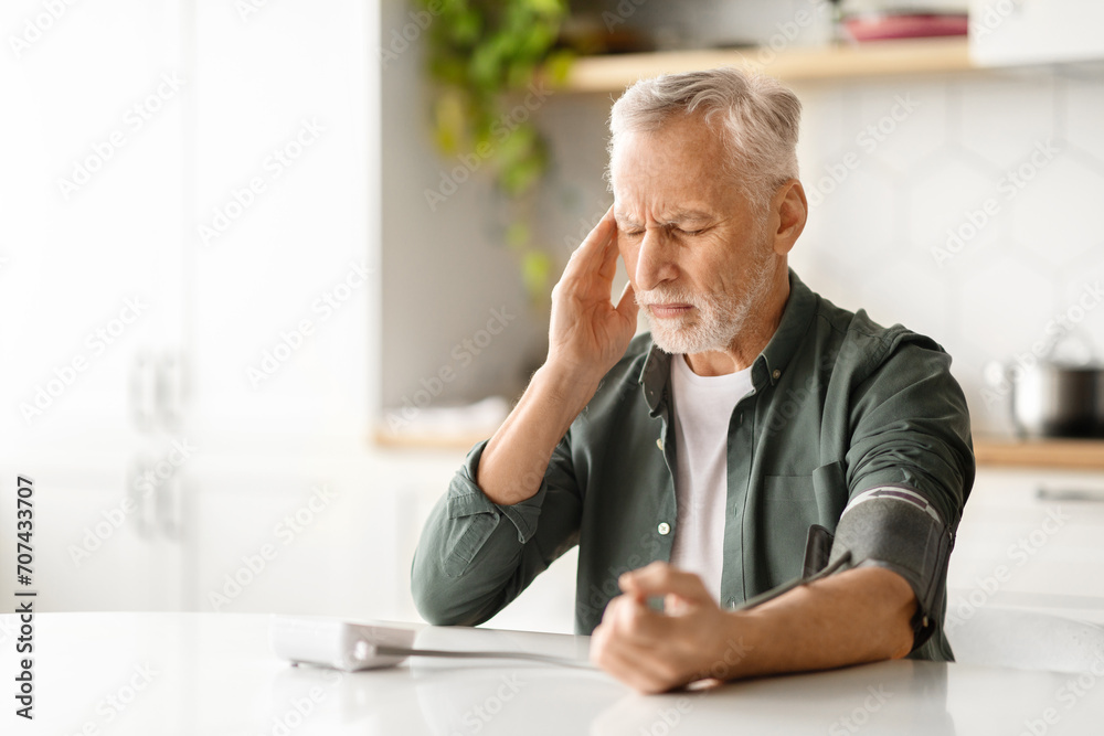 Senior man using upper arm blood pressure monitor while sitting at kitchen Senior man using upper arm blood pressure monitor while sitting at kitchen