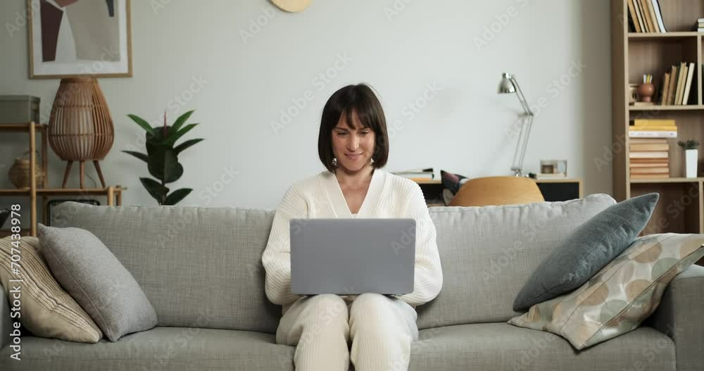 Cheerful woman sits on the couch, typing on laptop with a warm smile. Her positive demeanor and focused work ethic create an atmosphere of productivity and contentment in home.