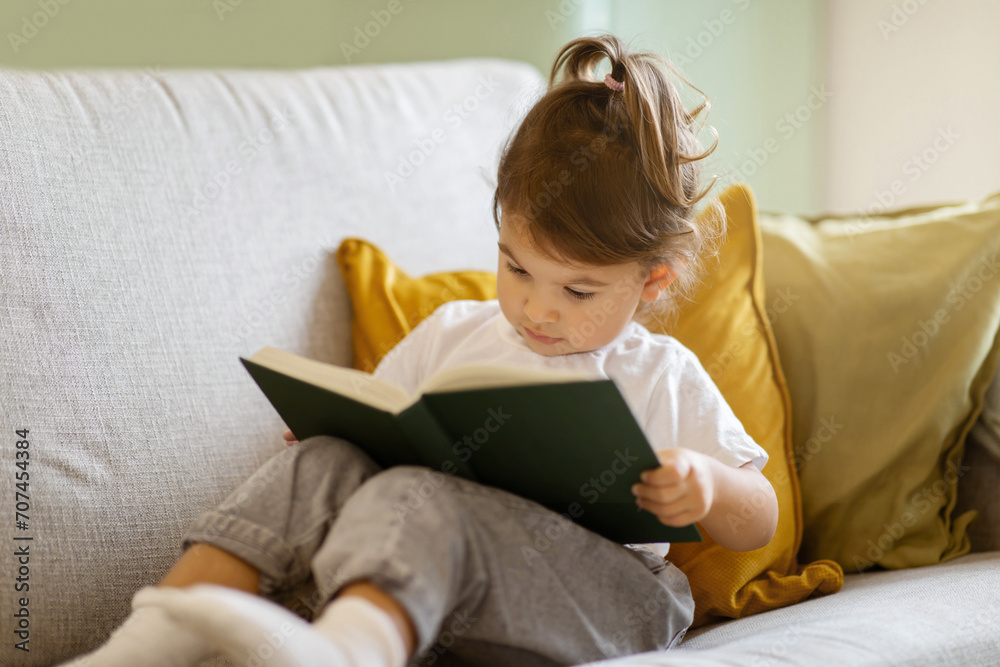 Cute little girl reading book while sitting on couch at home Cute little girl reading book while sitting on couch at home