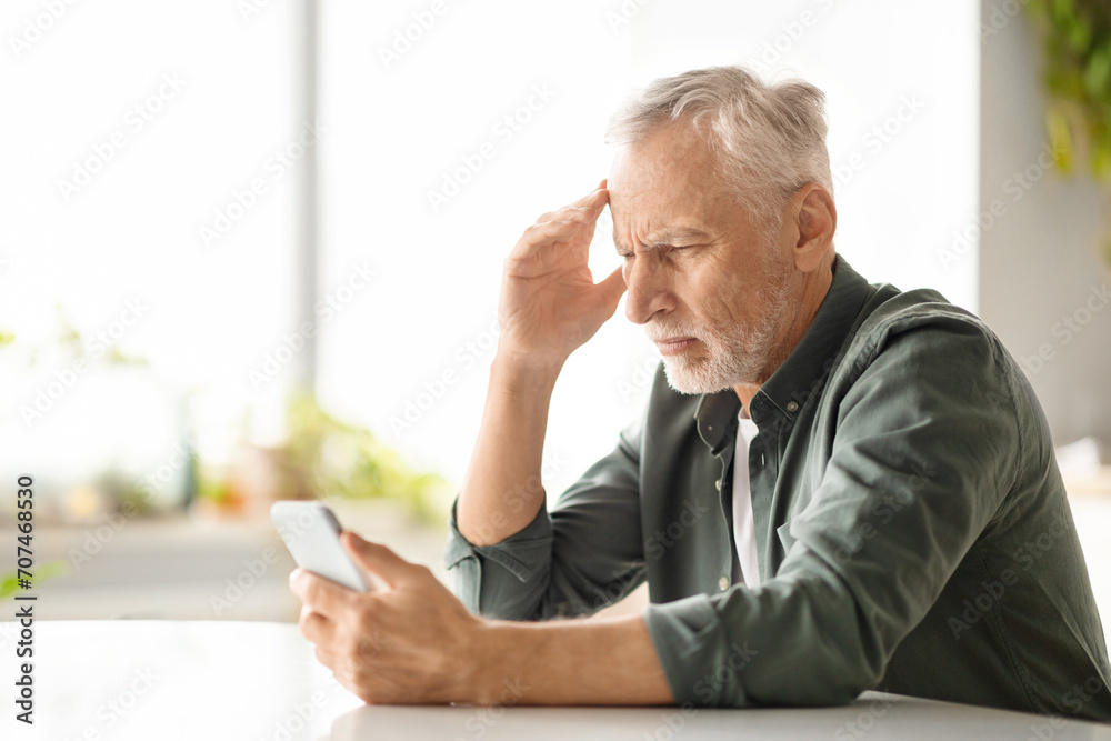 Distressed elderly man suffering headache while looking at smartphone at home Distressed elderly man suffering headache while looking at smartphone at home