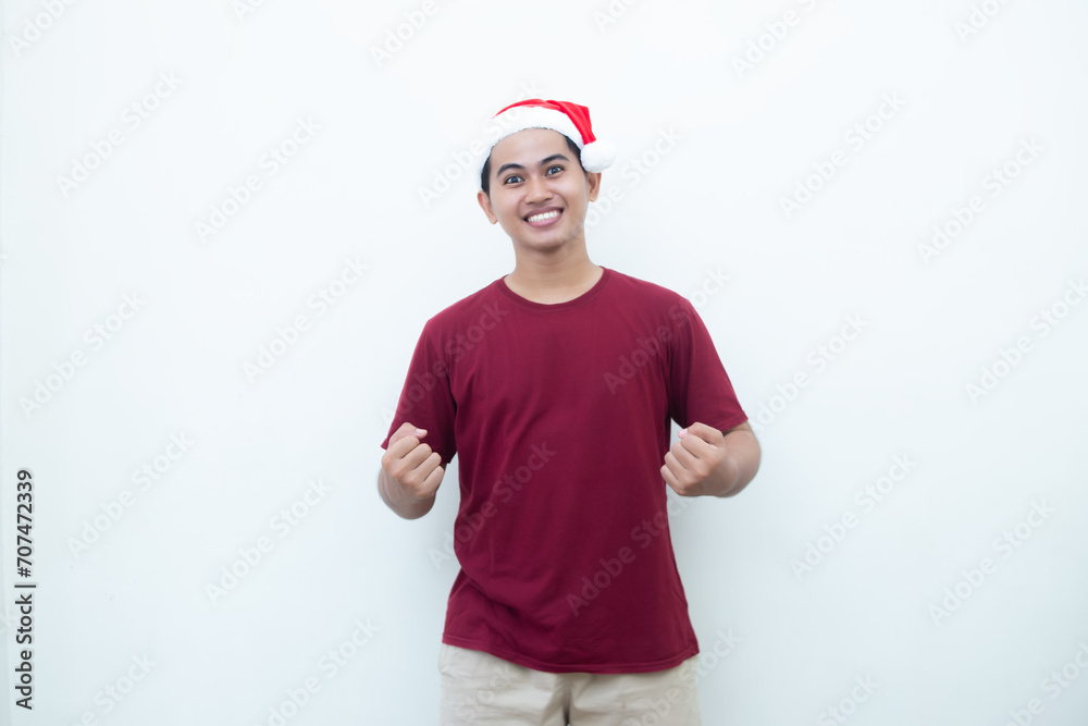 Young Asian man wearing a Santa Claus hat gripping both hands with enthusiasm and anger isolated by a white background for visual communication