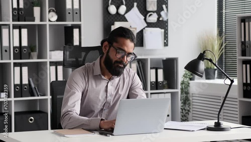 Indian businessman works on laptop in a bustling office environment. Arrayed in office appropriate clothing, he navigates through tasks with a focused expression, showcasing commitment to success. photo