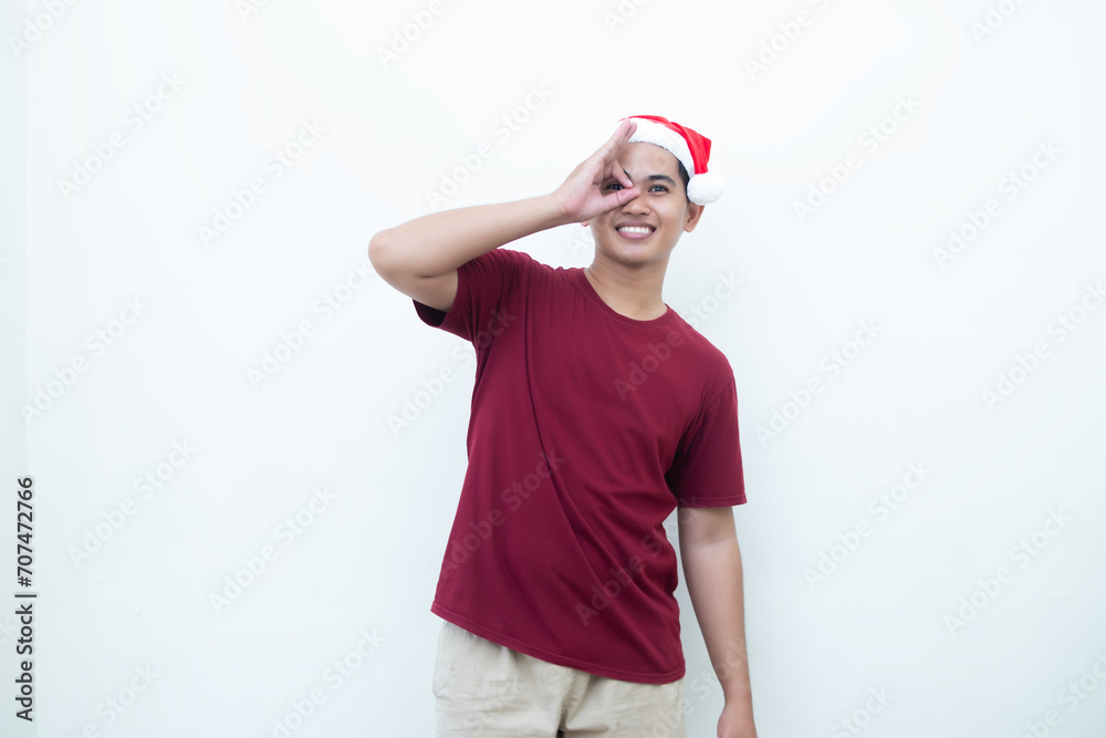 Young Asian man wearing a Santa Claus hat with a hand making the ok gesture isolated by a white background for visual communication Young Asian man wearing a Santa Claus hat with a hand making the ok gesture isolated by a white background for visual communication
