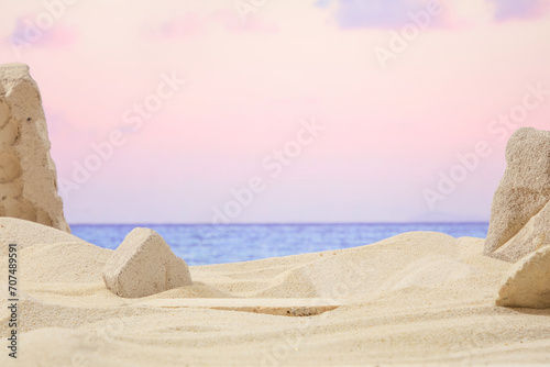 Beige sand background with surrounding gray rocks, in the distance is a pink sky with sunset appearing. Natural scene with empty space for design and display product