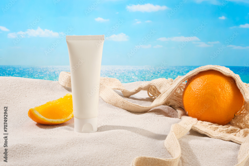 A tube of cosmetics without a label, an orange placed in a mesh basket on a white sand surface. Simulated beach scene for seasonal cosmetics advertisement. A tube of cosmetics without a label, an orange placed in a mesh basket on a white sand surface. Simulated beach scene for seasonal cosmetics advertisement.