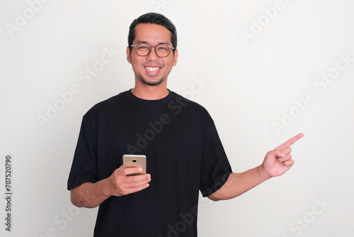 Adult Asian man smiling while holding phone with one hand pointing to the left side photo