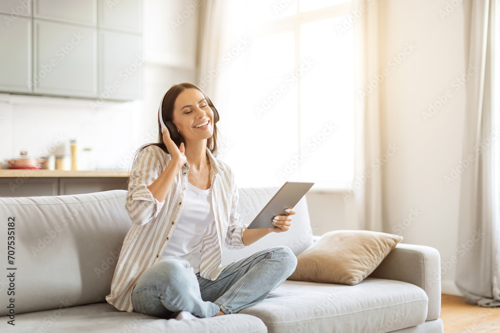 Joyful woman sitting cross-legged on a sofa, listening music through wireless headphones Joyful woman sitting cross-legged on a sofa, listening music through wireless headphones