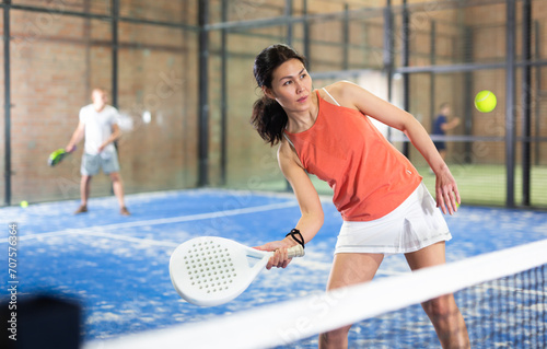 Portrait of concentrated asian woman playing paddle tennis indoors, preparing to hit forehand to return ball. © JackF