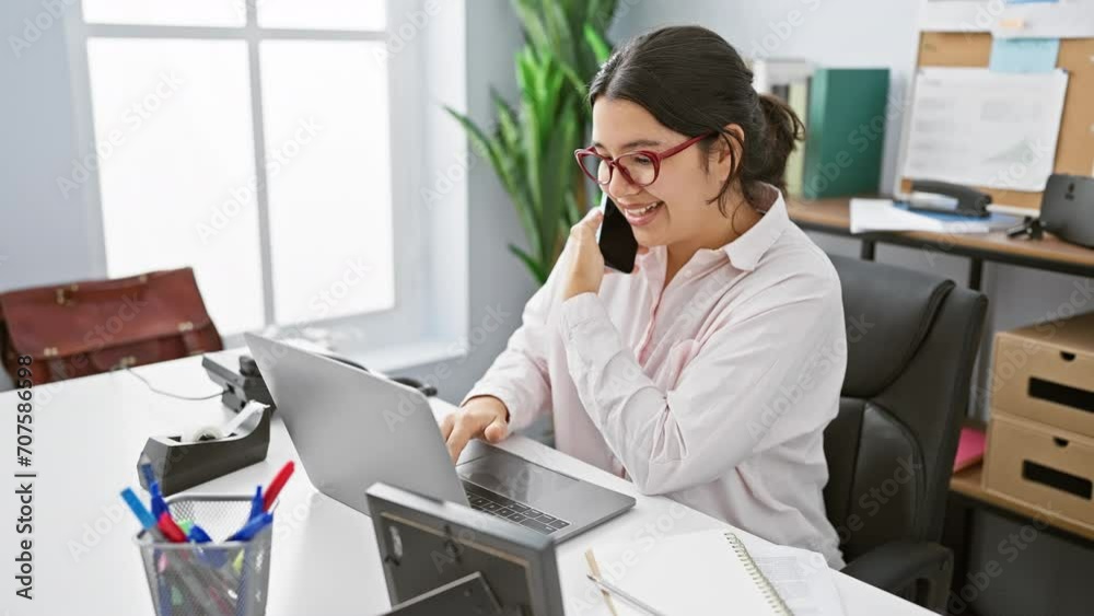 A young hispanic businesswoman multitasks in an office setting, smiling while on the phone and operating a laptop.