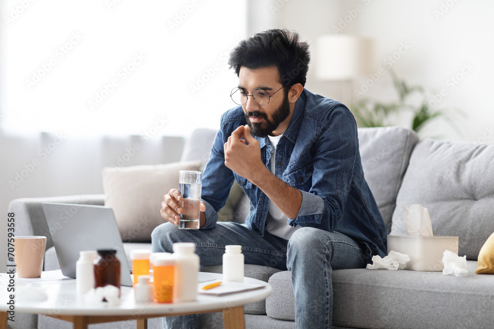 Ill indian man taking pill with water and looking at laptop screen - obrazy, fototapety, plakaty 