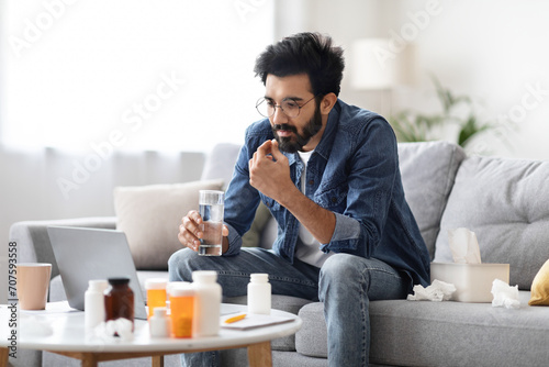 Ill indian man taking pill with water and looking at laptop screen photo