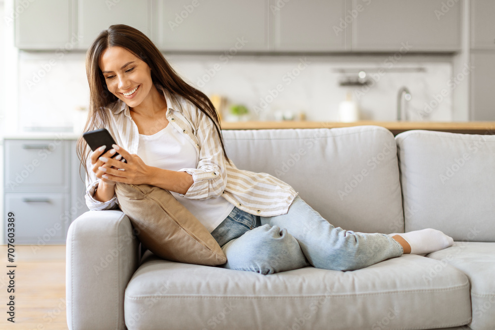 Contented female enjoying browsing her smartphone, sitting on couch in living room Contented female enjoying browsing her smartphone, sitting on couch in living room