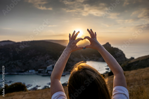 Happy woman on mountain peak, she makes heart shape with hands. Mountain, overlooking sea at sunset. Depicting love shape amidst scenic natural setting.