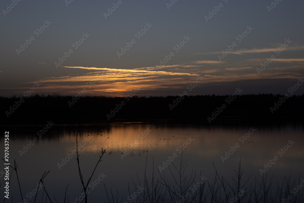 This image captures the serene mood of twilight over a still lake, where the silhouette of a forest horizon is set against a softly glowing sky. The dying light of the day peeks through the cloud