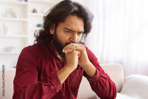 Sad bearded indian man sitting on couch at home