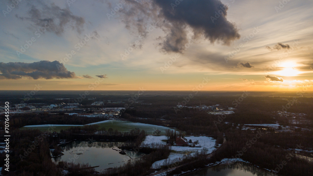 This aerial snapshot captures the serene ambiance of a winter evening as the sun sets, casting a warm glow that contrasts with the cool shadows of the day's end. The landscape, partially covered in