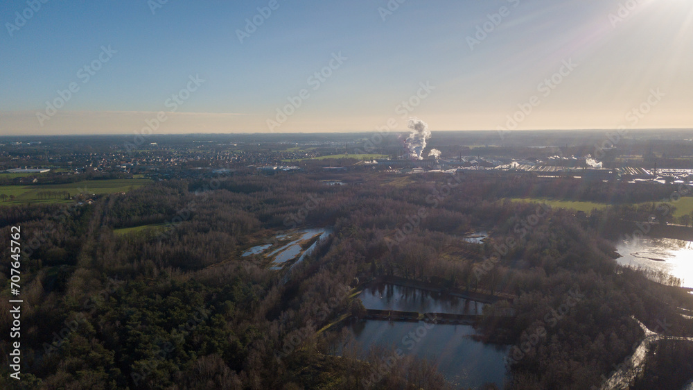 This aerial photo contrasts the tranquil beauty of a natural landscape with the distant bustle of industrial activity. In the foreground, serene bodies of water are surrounded by dense woodlands