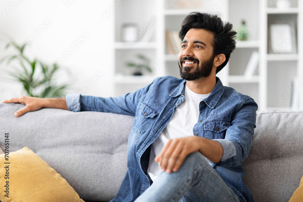 Handsome young indian man in denim shirt lounging on gray sofa at home