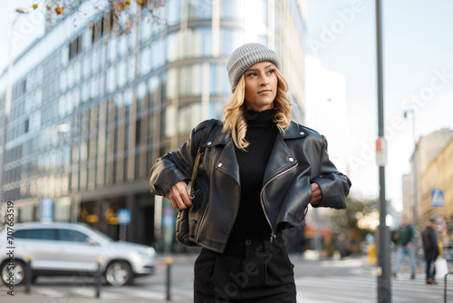 Stylish beautiful young woman in fashion urban clothes with a knitted hat, black sweatshirt, leather rock jacket walks in the city