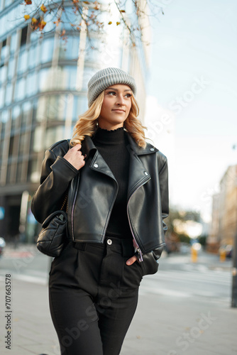 Urban stylish girl model with a knitted hat in a black sweater, leather jacket and trousers with a bag walks in the city
