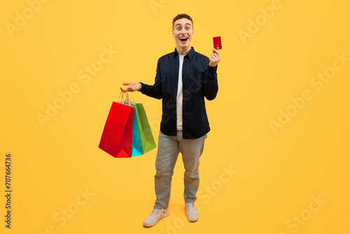 Excited young guy shopaholic holding purchases and credit card