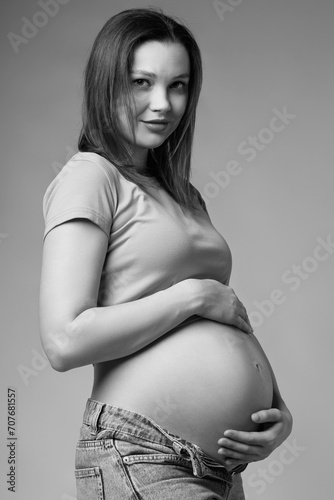Black and white portrait of young smiling pregnant woman in t-shirt and jeans on gray background.