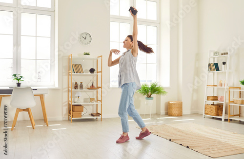 Full body photo of a young happy smiling girl wearing casual clothes dancing with smartphone in hand in the living room at home. Beautiful woman having fun with a satisfied face expression.