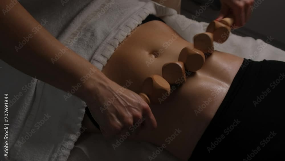 Woman performing anti-cellulite massage session with madero therapy on stomach, professional therapist holding wooden roller in studio.