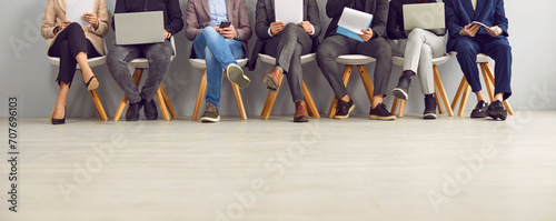 Group of people sitting in row in the office, waiting for a business meeting or job interview, working on laptop notebook PCs, holding clipboards. Cropped shot, human legs, banner background #707696103