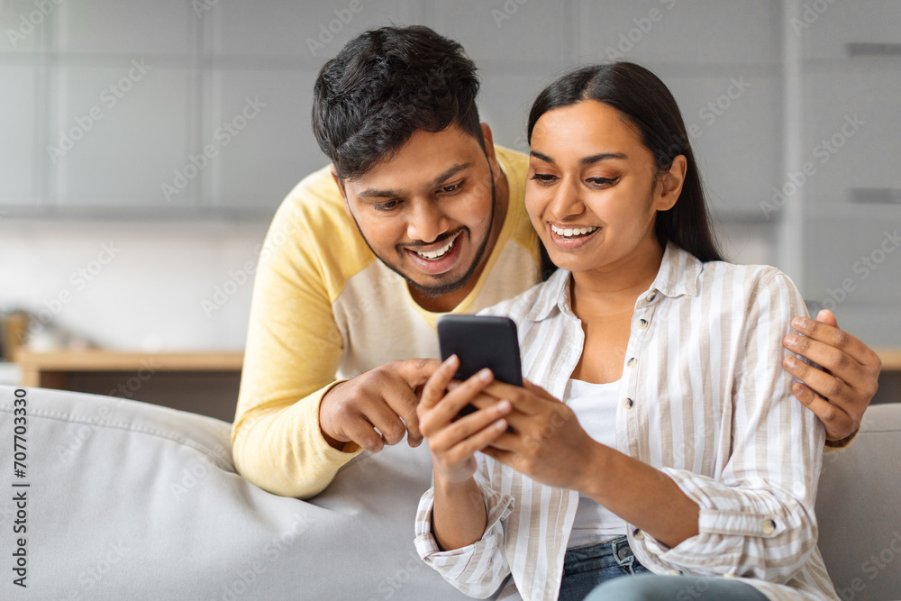 Cheerful Young Indian Couple Using Mobile Phone And Embracing Together At Home