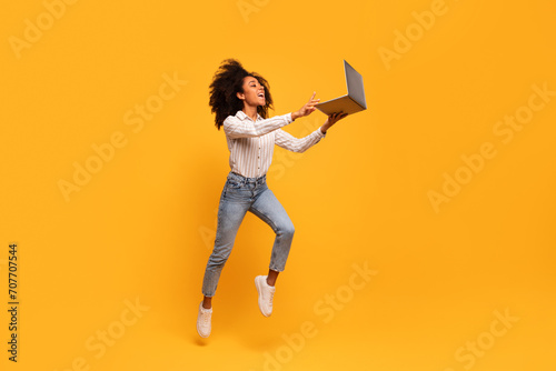 Foto Energetic black woman leaping with laptop on yellow background