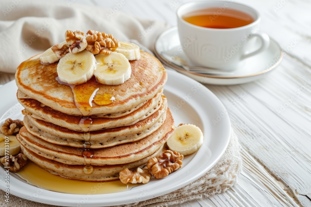 A white plate topped with banana and walnut pancakes, a cup of tea on the side, all served on a white wooden table.