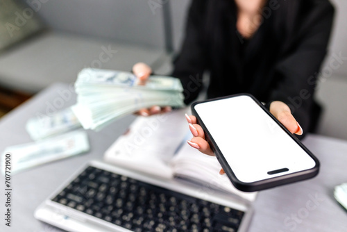 A woman's hand holds a black mobile phone with a white screen on an isolated background and money. Concept of financial services and modern technologies. Finance, transfers, banking services.