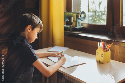 Boy is writing a letter at the table in his room in front of the window photo