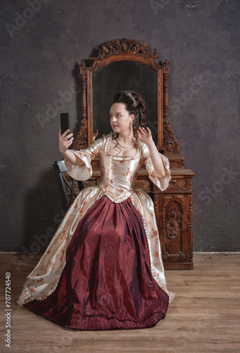 Beautiful woman in rococo style medieval dress sitting near console mirror table with mobile phone making selfie photo eclecticism concept