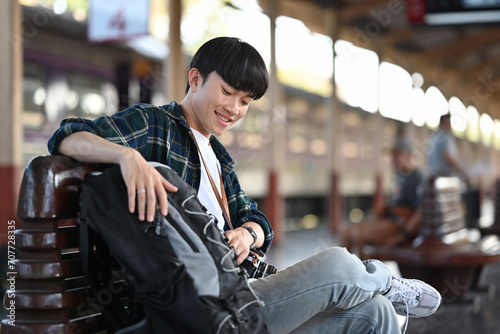 Young man backpacker sitting on a bench waiting train at railway platform. Travel and vacations concept