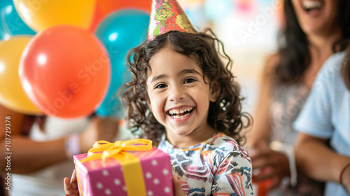 A child opening a birthday present with a huge smile surrounded by family and balloons.
