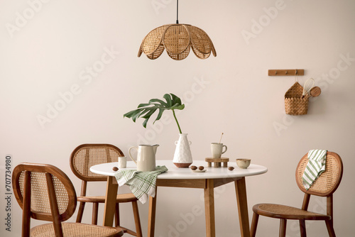 The stylish dining room with round table, rattan chair, lamp and kitchen accessories. Green leaf in vase.  Beige wall. Home decor. Template. photo