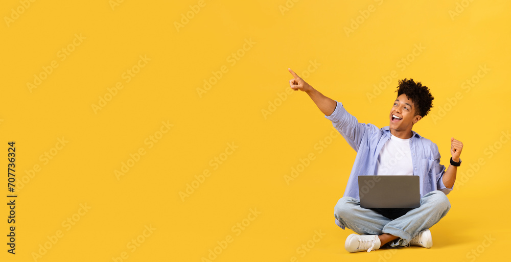 Joyful male student with laptop pointing at free space, sitting on yellow background Joyful male student with laptop pointing at free space, sitting on yellow background