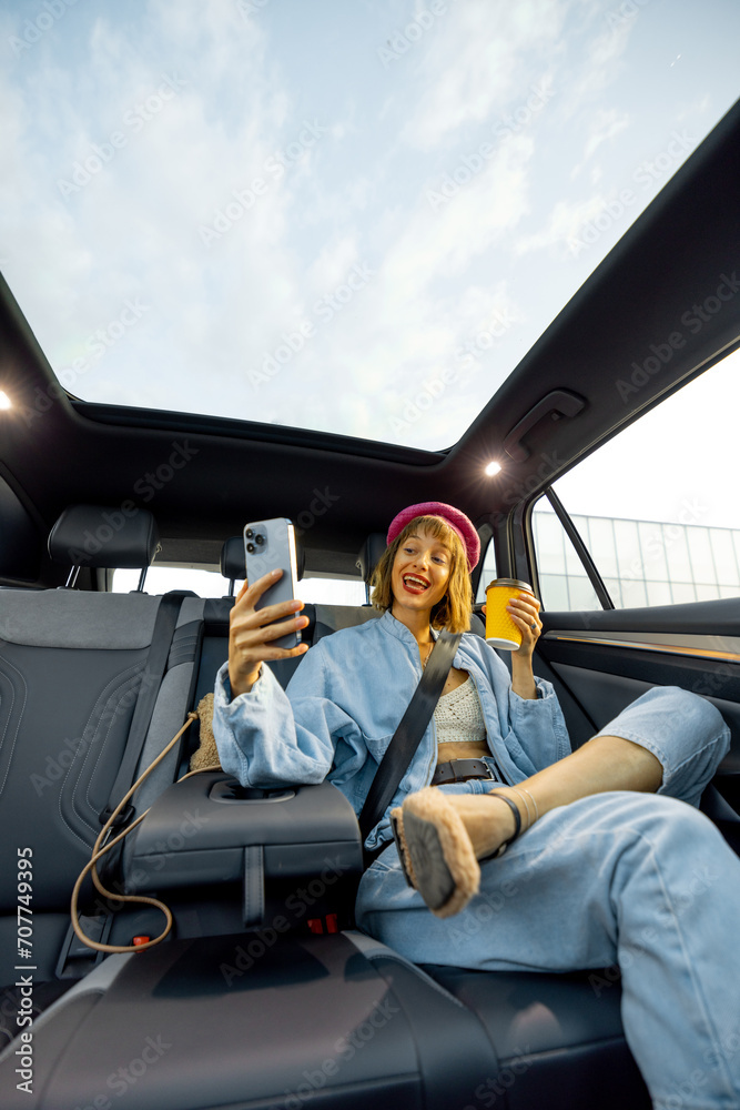 Young stylish woman uses smart phone while sitting relaxed with a coffee cup on backseat of car on the go. Wide angle view, modern car with panoramic rooftop