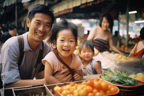A delighted Asian father shares a laugh with his young daughters  son and wife in the background  amidst colorful fruit stalls in a busy market. 
