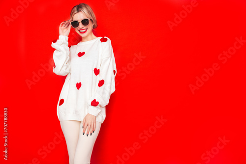 Young beautiful smiling female in trendy white sweater with hearts. Carefree woman posing near red wall in studio. Positive model having fun, going crazy. Cheerful and happy. Isolated